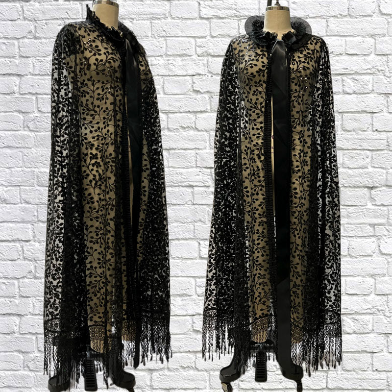 Ophelia sheer cape made of black sequin lace with lace fringe at the hem and ribbon detailing the neck and outer layer. A oversized black ribbon ties up at the neck. 