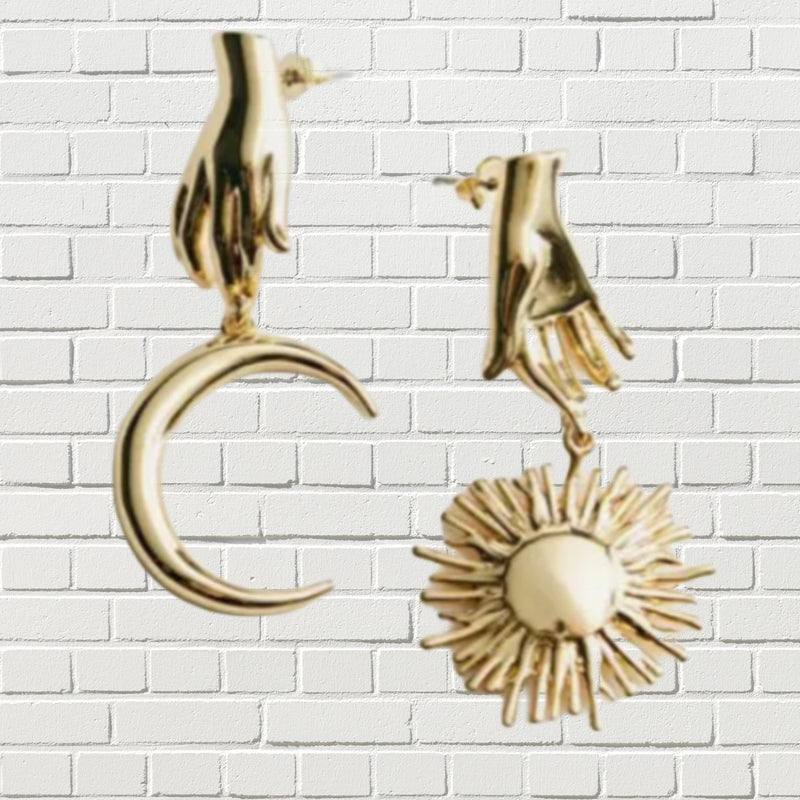 Gold plated celestial hands with moon and sun charms against a white brick backdrop.