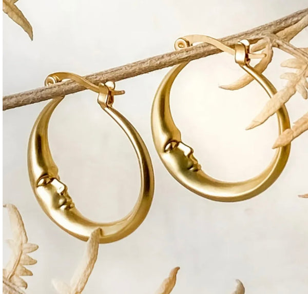 Crescent moon with a face earrings in gold hanging on a branch against a white backdrop.