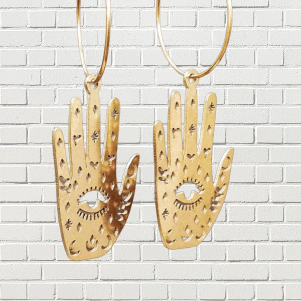 Delicate hoops with hanging mystic palm reading hands in bright gold against a white brick backdrop.