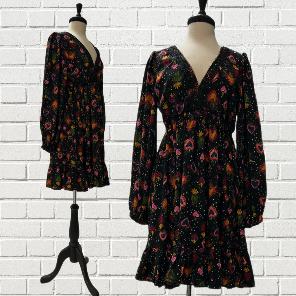 Mannequin form showing the front and side of the Francesca Baby Doll Dress in black with warm-toned sacred hearts, deep V-neck  with gimp lace trim, bishop sleeves, gathered empire waist, and a ruffled hem. 