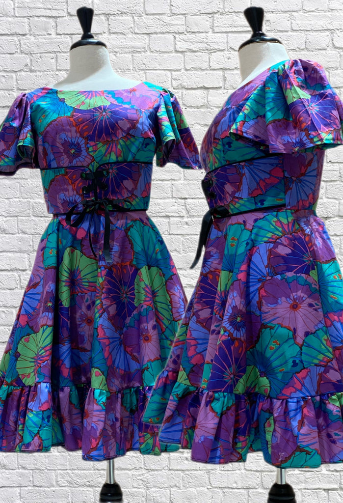 Mannequin form showing the front and side profile of the Purple and Teal floral cotton Jada dress, which has puff flutter sleeves, high neck fitted bodice with attached belt, ribbon tie up, circle skirt with ruffle hem, scooped back and side seam pockets.