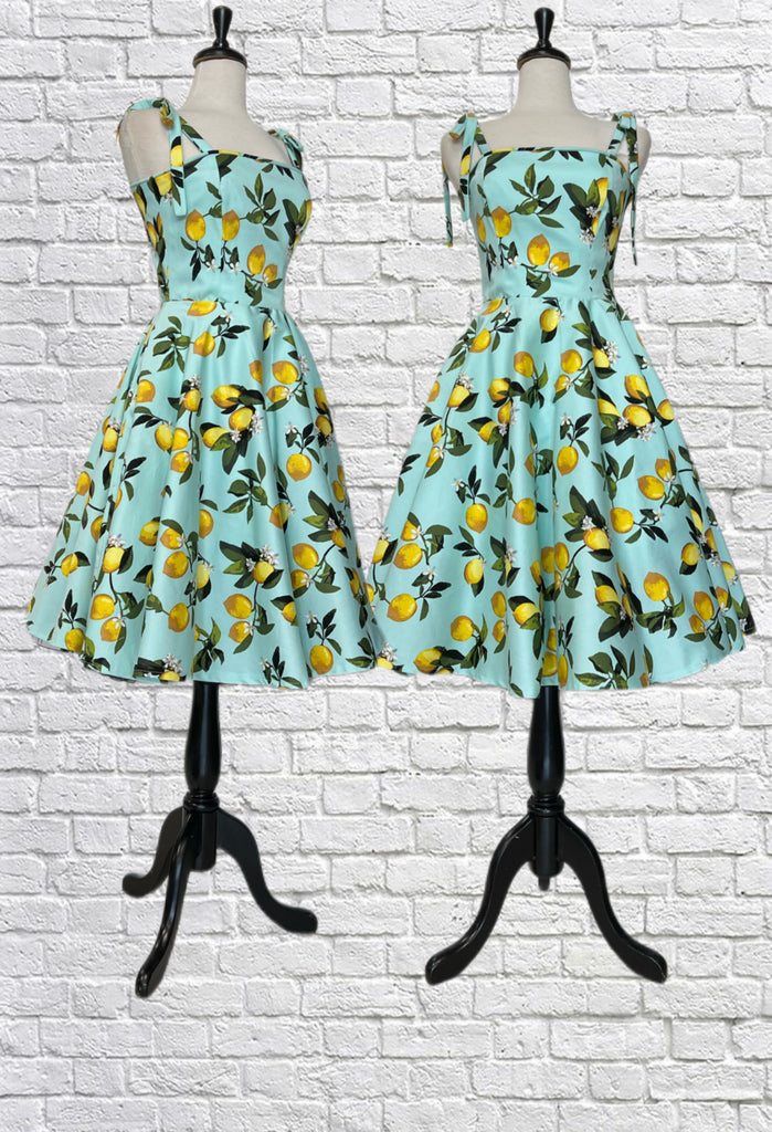 Mannequin forms showing the front of the Laura Dress in the perfect summer lemon print with a fitted bodice with adjustable bow straps, a circle skirt with full lining, and a back zipper closure.
