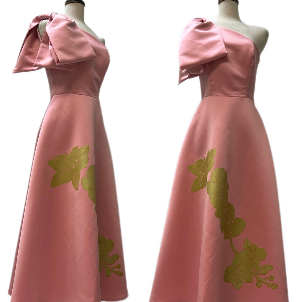 Two mannequin forms showing the side and front view of the Katie gown in blush pink Peau du Soie satin with fitted cold shoulder bodice, oversized shoulder bow, and A-Line maxi skirt with hand screen printed Orchid in gold. 